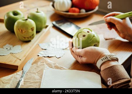 Idee di frutta fai da te di Halloween. Le mani femminili tagliano la mela verde di Halloween con la faccia creepy intagliata sul tavolo di legno nel paese. Spuntini salutari di Halloween Foto Stock