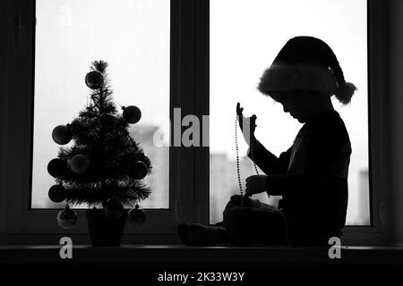 Silhouette bianca e nera di un ragazzo in costume da aiutante di Babbo Natale vicino a un albero di Natale Foto Stock