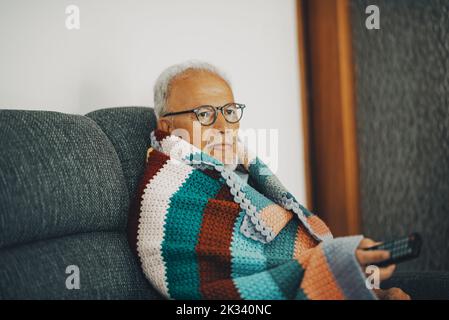 Un vecchio uomo anziano seduto sul divano a casa con copertura di lana colorata per i sintomi di bassa temperatura e influenza fredda. Concetto di crisi del gas energetico. Risparmio Foto Stock