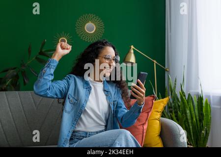 Felice e sorridente donna ispanica a casa è felice e sorridente leggere il messaggio online da smartphone, tenendo mano sul gesto di vittoria, celebrando il trionfo e il successo Foto Stock