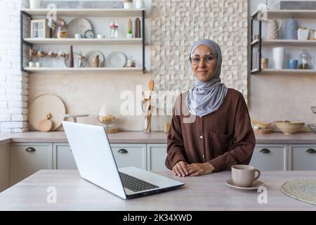 Ritratto di giovane donna musulmana in hijab, donna in occhiali guardando la fotocamera studiare a casa seduta in cucina, studente femmina utilizzando un computer portatile per l'apprendimento remoto on-line. Foto Stock