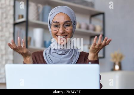 Videochiama online incontro con amici, giovane bella donna musulmana in hijab guardando la fotocamera e sorridendo parlando con gli amici a distanza, donna a casa in cucina utilizzando un computer portatile. Foto Stock