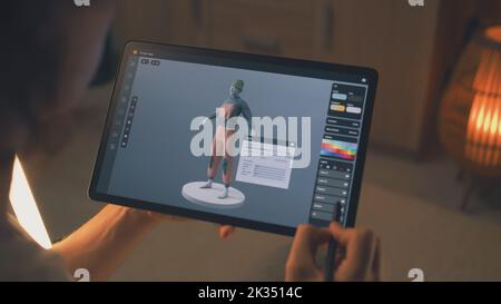 Designer 3D maschile che crea design di abiti in un'applicazione di modellazione 3D utilizzando computer tablet e matita digitale mentre lavora in remoto su un progetto freelance Foto Stock