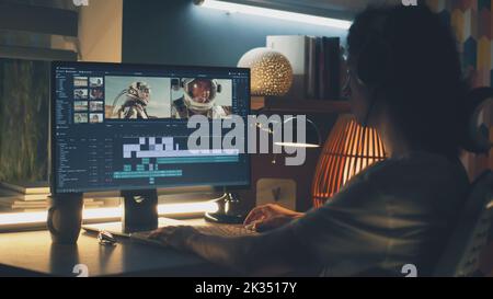 Editor maschile in cuffie seduti al tavolo di notte e editing di video con astronauti per i clienti su pc in programma professionale mentre lavorano in remoto Foto Stock
