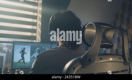 Vista posteriore di un uomo in cuffie seduto al tavolo e la creazione di eroe videogiochi e animazione 3D in un programma professionale per computer, mentre si lavora da remoto a casa. Foto Stock