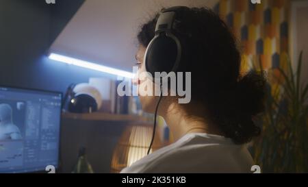 Vista posteriore di un uomo in cuffie seduto al tavolo e la creazione di eroe videogiochi e animazione 3D in un programma professionale per computer, mentre si lavora da remoto a casa. Foto Stock