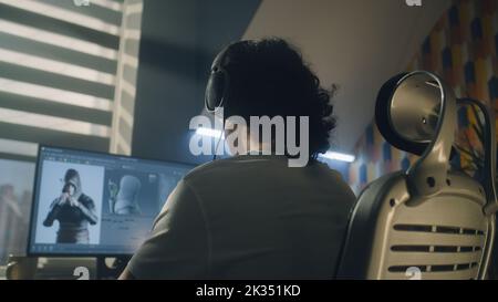 Vista posteriore di un uomo in cuffie seduto al tavolo e la creazione di eroe videogiochi e animazione 3D in un programma professionale per computer, mentre si lavora da remoto a casa. Foto Stock