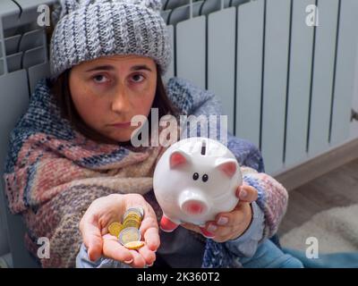 Donna che conta monete in euro e tiene il piggy bank vicino alla batteria a casa. Donna vestita calorosamente è sconvolta circa il prezzo elevato di riscaldamento. Costoso it Foto Stock
