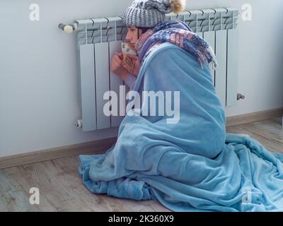 Donna seduta e contante banconote in euro vicino alla batteria a casa. Donna vestita calorosamente è sconvolta circa il prezzo elevato di riscaldamento. Energia costosa re Foto Stock