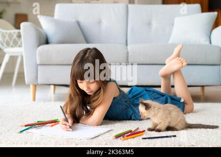 Una ragazza simpatica e divertente in una sundress denim e scopa nei suoi capelli si trova in una stanza su un tappeto e disegna con matite colorate. Accanto a lei c'è un fr a quattro zampe Foto Stock