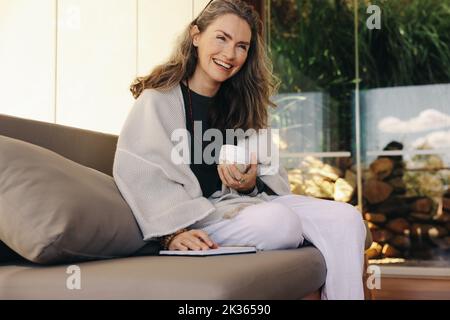 Allegra donna anziana che giornalava con una tazza di tè al mattino. Donna matura sorridente felice mentre si siede su un divano. Donna anziana che si gode relaxi Foto Stock