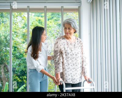 Caregiver, sorridente giovane asiatica femmina, figlia o nipote aiutare donna anziana, madre o nonni cercare di camminare con la struttura del camminatore a h Foto Stock