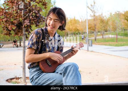 Ragazzo taiwanese sorridente e suonante la chitarra acustica Ukulele seduta su una panchina. Foto Stock