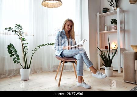 Giovane donna in tuta che legge un libro seduto su una sedia Foto Stock