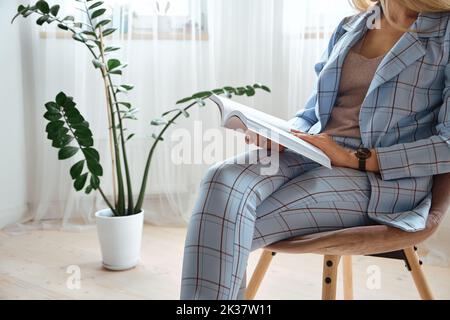Irriconoscibile giovane donna in tuta che legge un libro seduto su una sedia Foto Stock