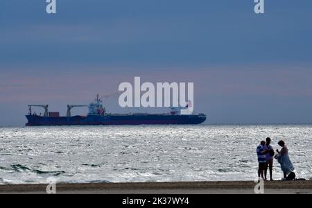 Tre bagnanti chiacchierano sulla spiaggia al tramonto con una grande nave portacontainer all'orizzonte. Foto Stock