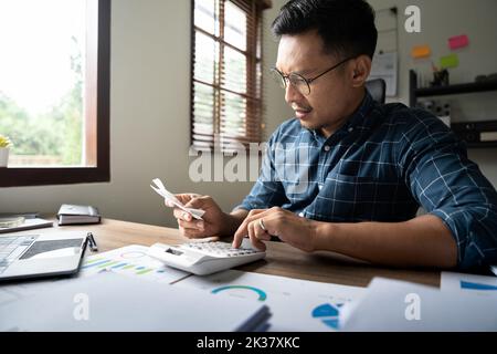 Il giovane asiatico che calcola le spese della famiglia circa la finanza ed è frustrato sulla scrivania nel paese, il controllo maschio che legge ha lo sforzo e preoccupato, il debito e. Foto Stock