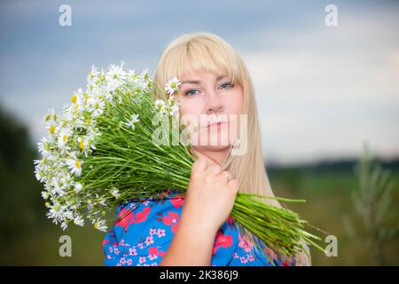 Una bella ragazza più taglia con capelli bianchi in un abito estivo posa sulla strada con margherite. Chubby ragazza in un prato con margherite primo piano. Foto Stock