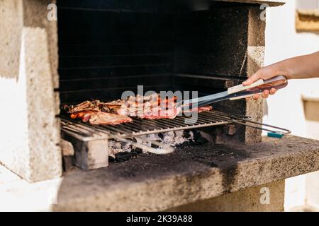 mani di un uomo caucasico che gira un filetto di maiale su un barbecue con un paio di pinze Foto Stock