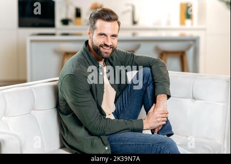 Foto di un bell'elegante uomo caucasico bearded, in abiti casual, seduto su un divano in un accogliente soggiorno, rilassarsi dopo una giornata di lavoro, guardando la macchina fotografica, sorridendo felicemente Foto Stock