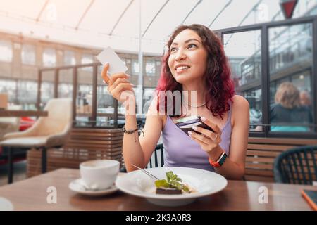 Una ragazza felice ha alzato la mano e chiama il cameriere per pagare la colazione al bar con una carta di credito Foto Stock