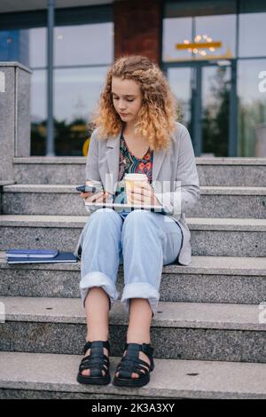 Ragazza studente è seduta sulle scale con il computer portatile in grembo, utilizzando i social media con smartphone che hanno una discussione interessante. Istruzione, scuola Foto Stock