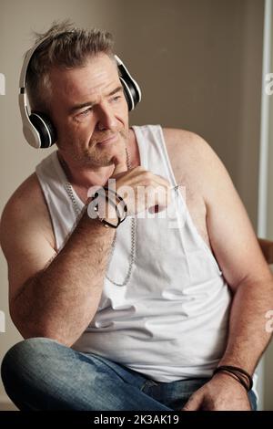 Uomo maturo sorridente in canotta bianca che ascolta la musica con le cuffie e guarda lontano Foto Stock