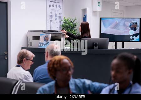 Addetto al ricevimento asiatico che lavora con la stampante presso la reception hopital per fare referti medici e moduli di regrazione per i pazienti con appuntamenti. Utilizzando carte per il supporto di assicurazione sanitaria. Foto Stock