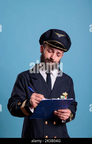 Pilota di aeroplano che tiene appunti, compila il modulo, aviatore concentrato con documenti. Capitano in uniforme in vista frontale dell'aeroporto, ripresa media da studio su sfondo blu Foto Stock