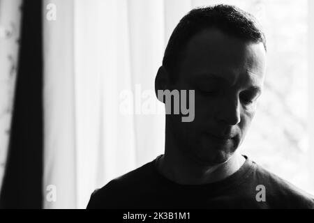 Ritratto di studio scuro di un giovane uomo caucasico adulto in piedi vicino a una luminosa finestra illuminata, foto artistiche in bianco e nero con messa a fuoco selettiva e morbida Foto Stock