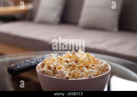 Ciotola con popcorn e telecomando tv su tavolo in vetro vicino al divano Foto Stock
