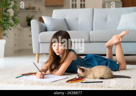 Una ragazza simpatica e divertente in una sundress denim e scopa nei suoi capelli si trova in una stanza su un tappeto e disegna con matite colorate. Accanto a lei c'è un amico a quattro zampe - un piccolo gattino divertente e soffice. Foto di alta qualità Foto Stock