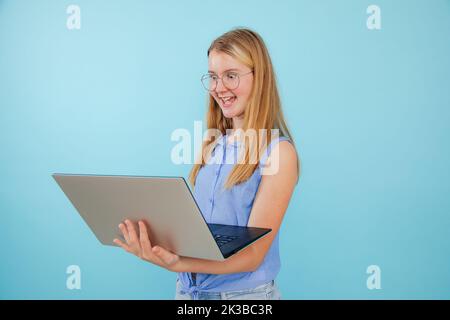 Ritratto di ragazza giovane stupita con capelli lunghi indossando camicia blu senza maniche, occhiali che tengono il laptop su sfondo blu. Foto Stock