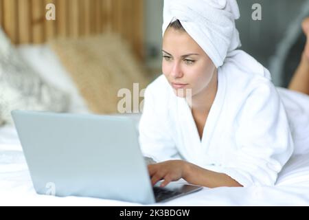 Bella donna sdraiata su un letto dopo aver fatto la doccia usando il notebook a casa Foto Stock