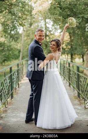 Felice coppia di nozze sorridente in piedi sul ponte nel parco in estate, guardando indietro. Giovane sposa che si prepara a lanciare bouquet. Foto Stock