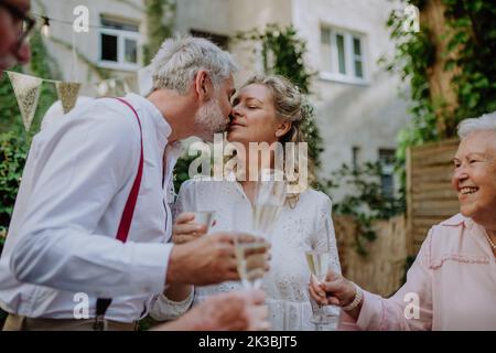 Sposa matura e sposo baciare al ricevimento di nozze con la loro famiglia, fuori nel cortile. Foto Stock