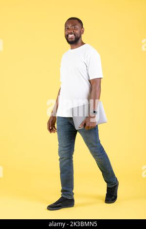 Allegro African American Guy Holding laptop che cammina su sfondo giallo Foto Stock