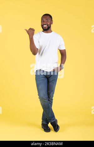 Vertical Shot of Cheerful Black Guy che punta il pollice da parte, Studio Foto Stock