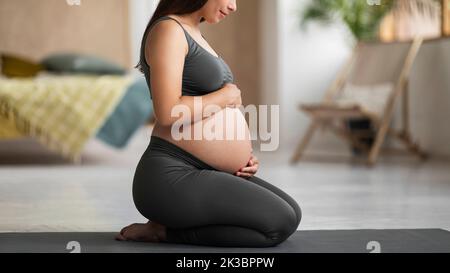 Giovane donna incinta che abbraccia il ventre mentre si siede sul tappetino Yoga a casa Foto Stock