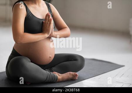 Donna incinta irriconoscibile in abbigliamento sportivo meditando a casa, primo piano colpo Foto Stock