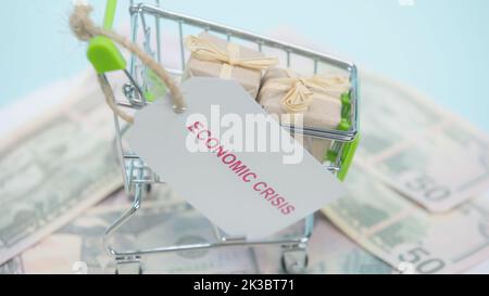 primo piano del carrello della spesa con regali e scheda con la scrittura di crisi economica sui dollari e sfondo blu, immagine di riserva Foto Stock