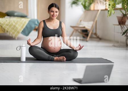 Donna incinta sorridente con il laptop che pratica lo yoga in linea a casa Foto Stock