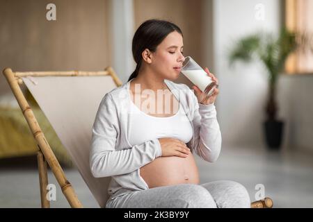 Donna incinta bere latte dal vetro mentre si rilassa in sedia a casa Foto Stock