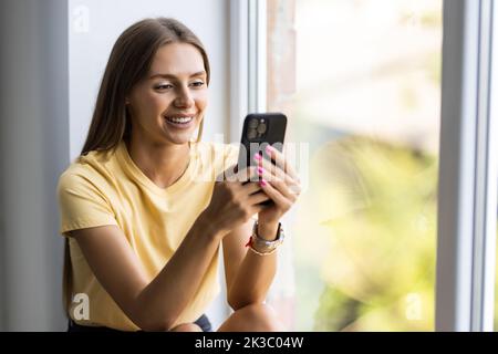 Attraente giovane donna seduta su un davanzale a casa e testando sul suo telefono cellulare comunicazione femmina guardando messaggio cellulare allegro sorriso utilizzare inte Foto Stock