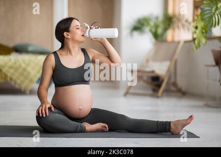 Giovane donna incinta bere acqua dalla bottiglia durante l'allenamento di yoga a casa Foto Stock