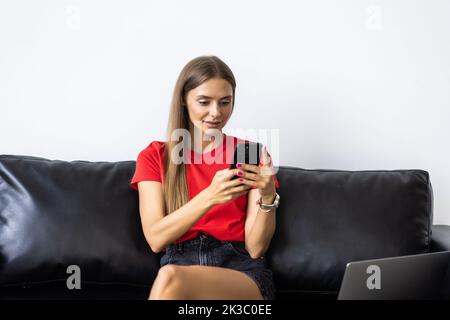 Affascinante, bella donna seduta sul divano in soggiorno a casa, facendo shopping online attraverso 3G internet, avendo smartphone in mano Foto Stock