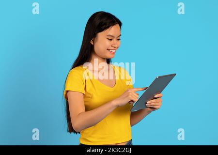 Felice occupato giovane studentessa coreana in t-shirt gialla chattando su tablet, isolato su sfondo blu, studio Foto Stock