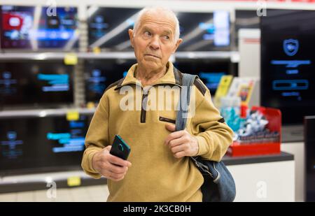 uomo anziano in grigio pensionato in cerca di bancone con moderni televisori digitali in showroom di digital goods store Foto Stock