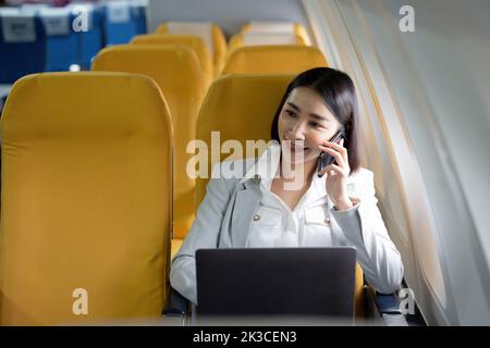 Allegra donna d'affari asiatica che chiama il telefono e usa il computer portatile durante il volo. Stile di vita urbano Foto Stock