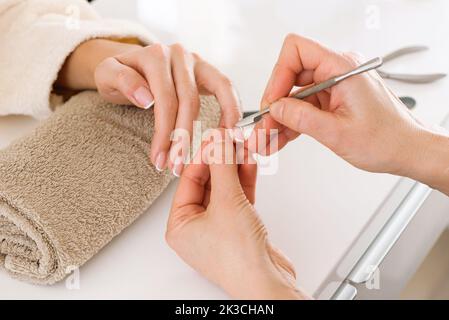 Da sopra il tecnico del chiodo che taglia la cuticola sul dito della donna che tiene la mano su asciugamano arrotolato su tabella bianca in salone di stazione termale Foto Stock